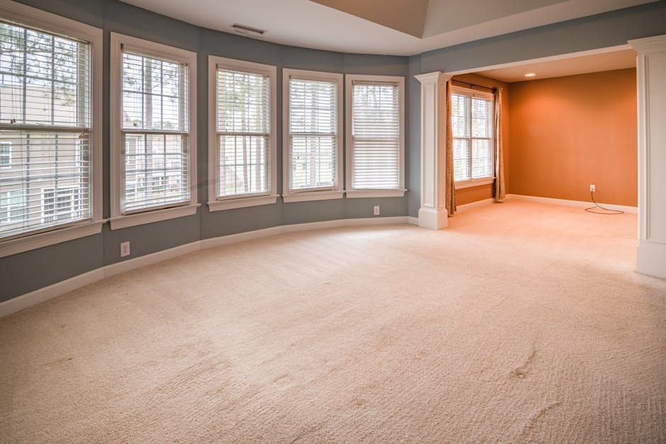 A woman with curly blonde hair, dressed in a beige jacket, jeans, and white shoes, is engaged in surface cleaning in a living room. She is vacuuming a patterned area rug with a yellow vacuum cleaner, 