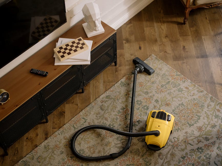 Close-up of a modern, upright vacuum cleaner with a transparent dust container filled with debris, positioned on a pink carpeted floor in a room. The vacuum has a flexible, patterned hose attached to